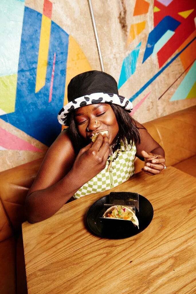 A young woman enjoying our halloumi tacos wearing a chequered top and cow print hat against a colourful wall mural