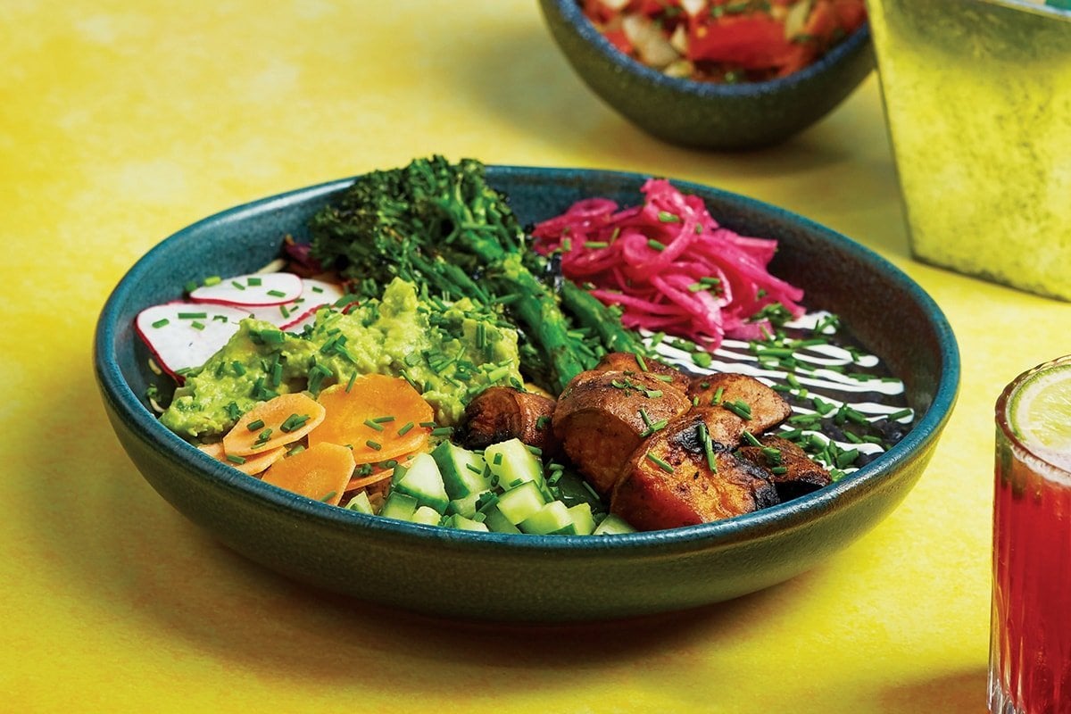Veggie rainbow bowl with sweet potato and broccoli, alongside frijoles, pink pickled onions, green rice, slaw and guacamole