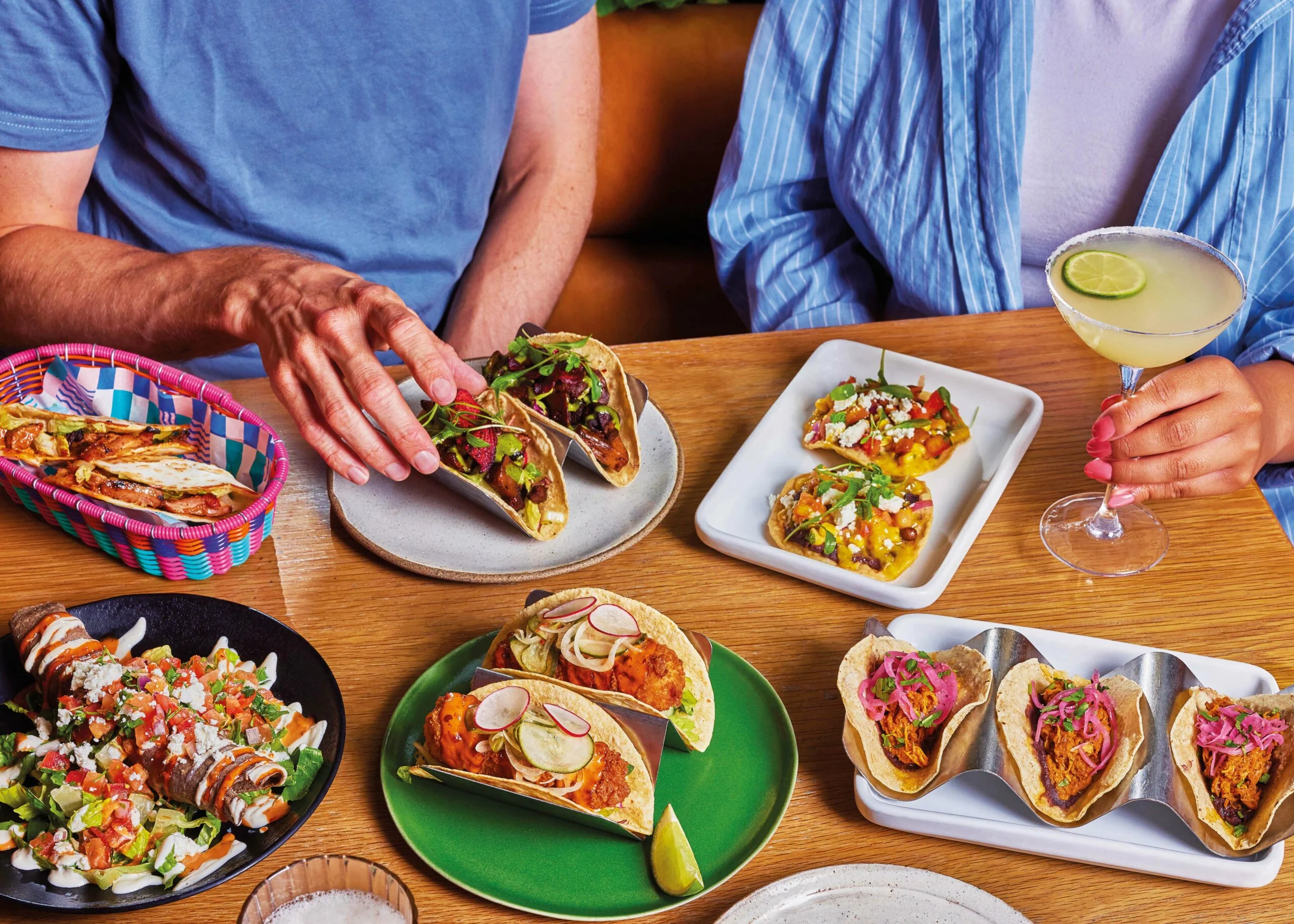 A feast of Wahaca plates on a table, with two people tucking in to tacos and a margarita