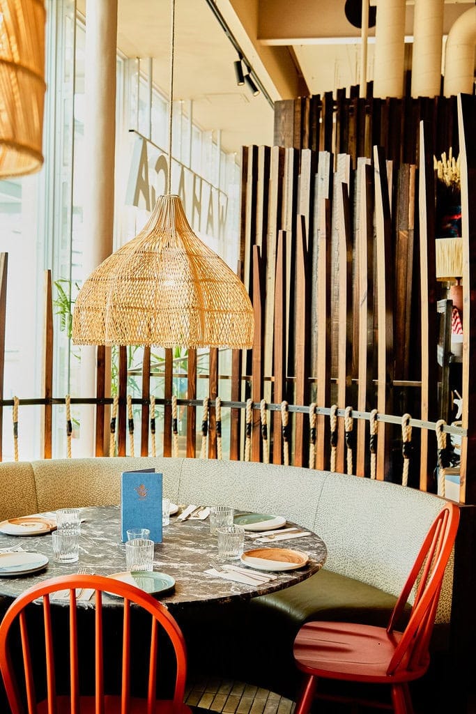 A booth table in our Mexican restaurant in London. Inspired by a treehouse, it is surrounded by wooden bars and has a low hanging straw lampshade hanging above the table to create an intimate feeling.