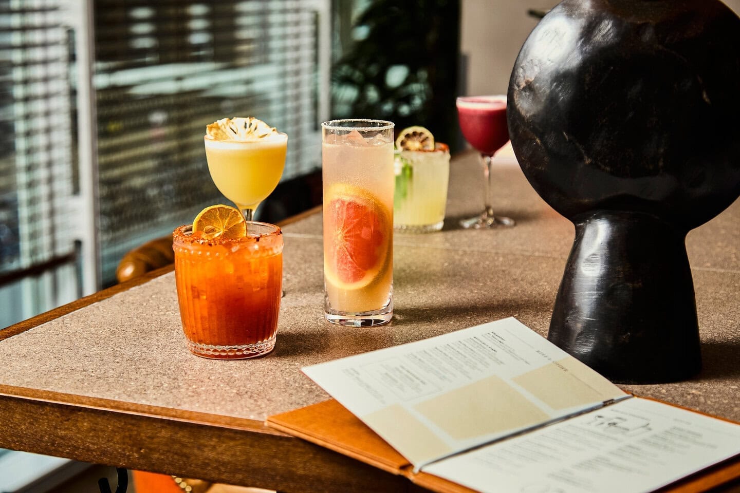 five freshly shaken and colourful cocktails sit on the bar of our mexican restaurant in london, they include an orange coloured marmalade mezcalita, a pink paloma, a yellow pineapple mezcalita, a purple blackberry sour and a picante margarita