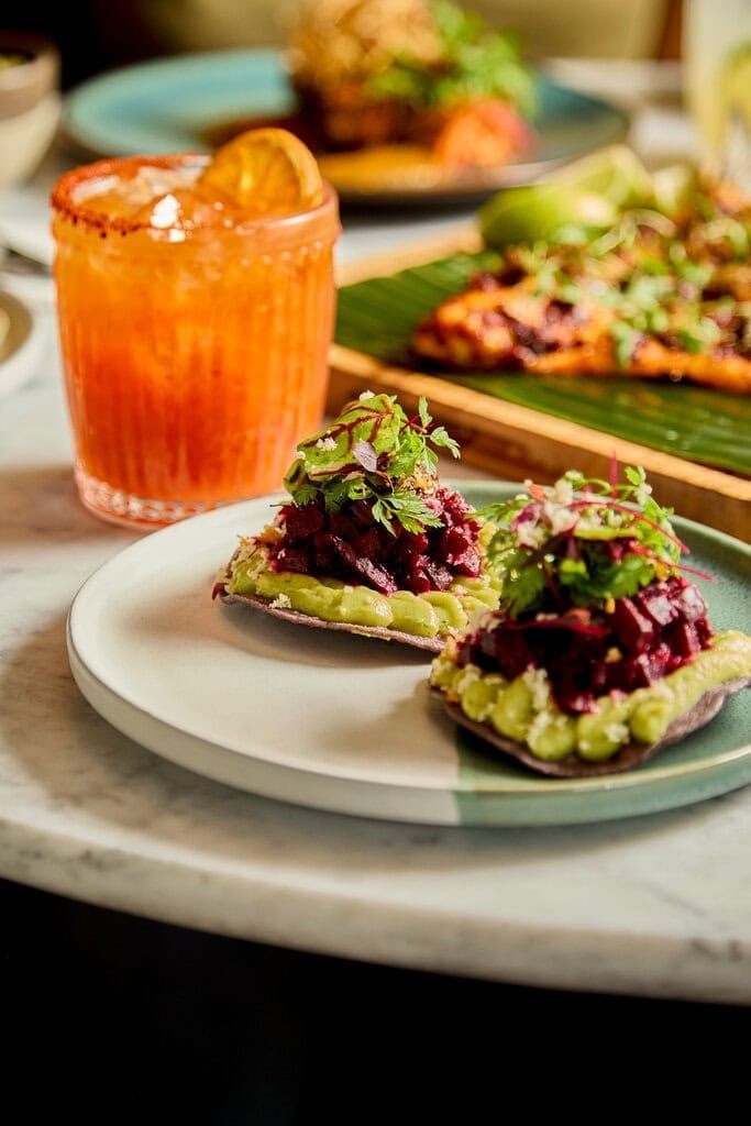 a table in a mexican restaurant contains a plate of mexican tostada topped with beetroot and avocado crema, a marmalade mezcalita cocktail and a achiote chilli marindade fish in the background.