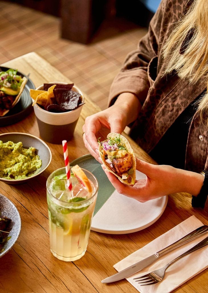 A woman sits at a table in our Mexican restaurant in London holding a vegetarian taco. She also has a non-alcoholic citrus drink and some fresh guacamole with tortilla chips.