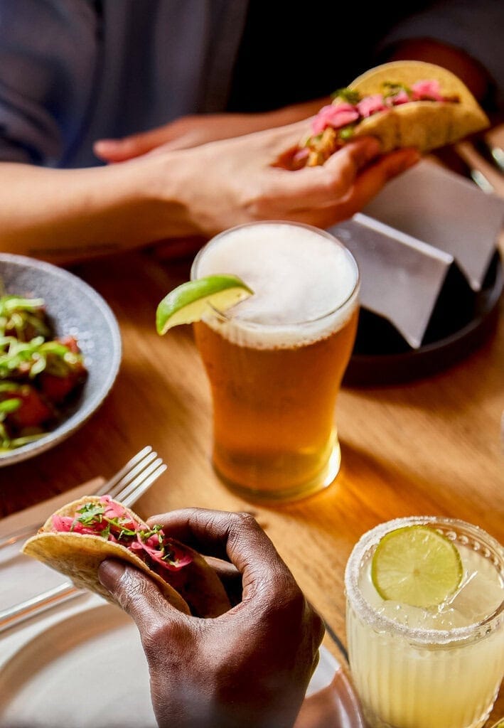 Two people hold a taco each - they both hold a pork pibil taco with pink pickled onions. A pint of lager beer sits on the table between them, which is garnished with a fresh lime slice. There is also a classic margarita cocktail on the table with a salt rim.