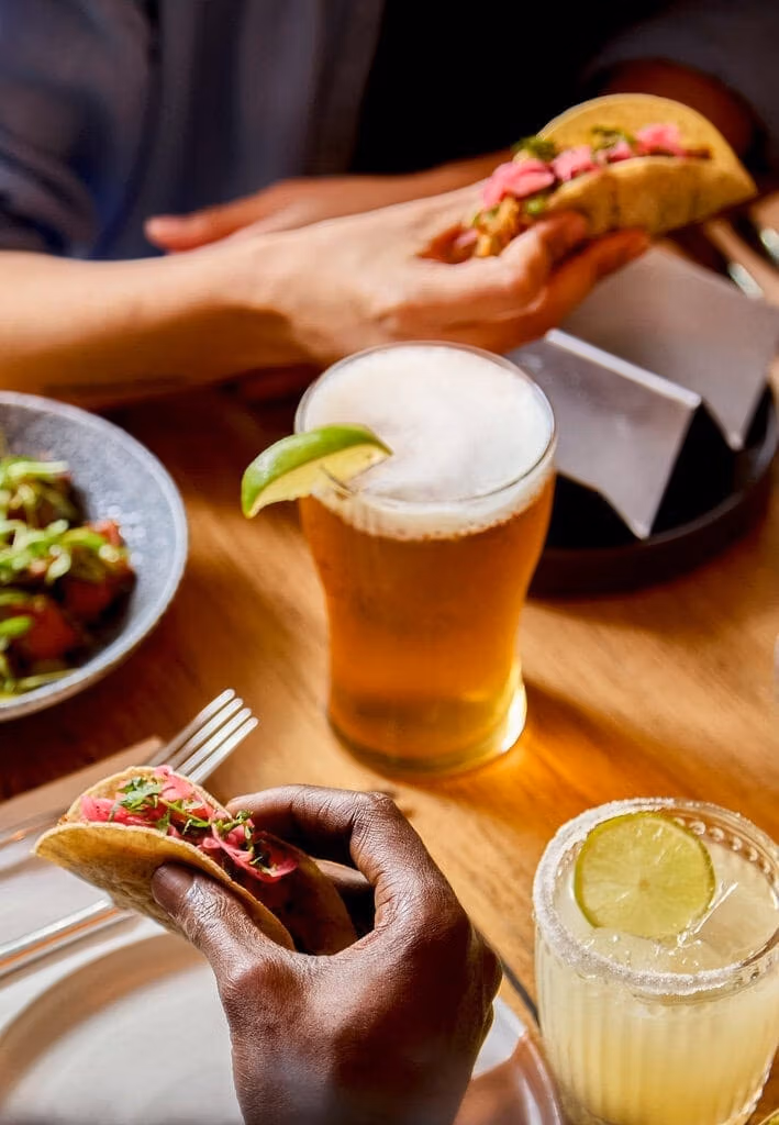 Two people hold a taco each - they both hold a pork pibil taco with pink pickled onions. A pint of lager beer sits on the table between them, which is garnished with a fresh lime slice. There is also a classic margarita cocktail on the table with a salt rim.